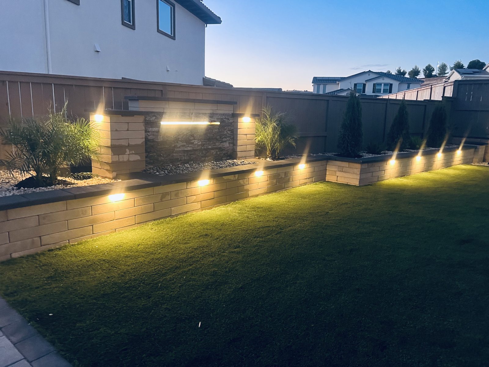 Garden lighting washes across the lawn and highlights the textured boundary wall.